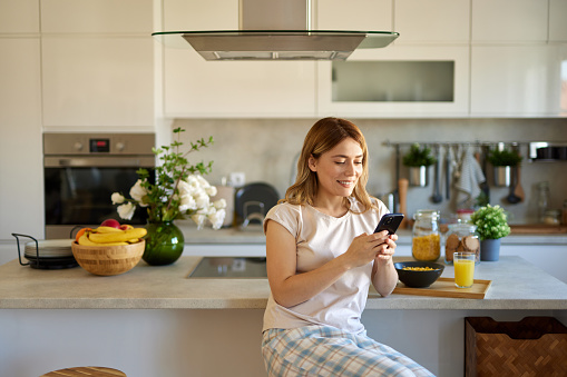 Portrait of a woman eating breakfast while using mobile phone in the kitchen