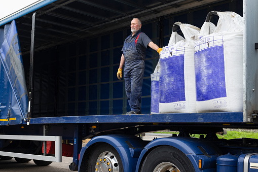 A shot of a delivery driver standing on the back of an articulated lorry delivering nitrogen sulphur fertiliser to a farm in Northumberland in northeast England. He is leaning against one of the bags.