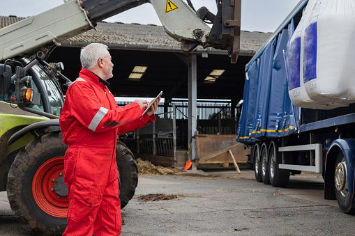 A shot of a male farmer standing using digital technology collecting data as he receives a delivery of nitrogen sulphur fertiliser. There is a forklift tractor lifting the delivery of fertiliser from an articulated lorry.