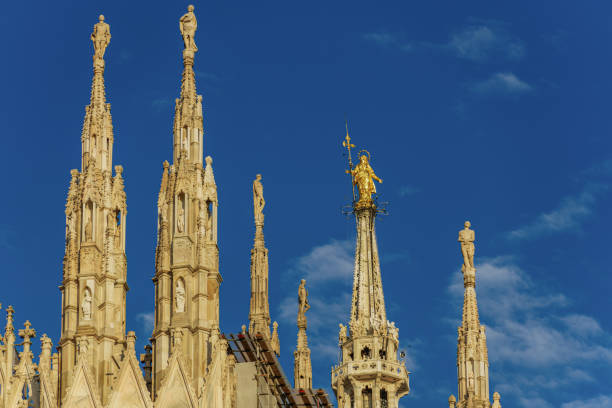 Milan, Italy day view of gothic style Roman Catholic Duomo Cathedral rooftops at homonym main square, with Statue of Madonnina, the Little Madonna, Virgin Mary golden artwork visible against blue sky. milán, italia, vista diurna de los tejados de la catedral católica romana del duomo de estilo gótico en la plaza principal homónima, con la estatua de la madonnina, la pequeña virgen, la obra de arte dorada de la virgen maría visible contra el cielo