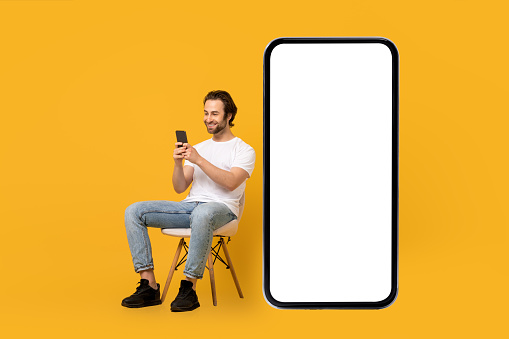 Smiling young european male in white t-shirt sits on chair, typing on phone near huge smartphone with empty screen isolated on yellow background, studio. Blog, chat, ad and offer, app and website