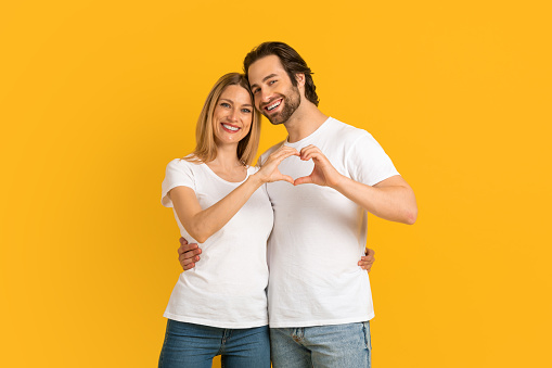 Happy attractive millennial european wife and husband with stubble in white t-shirts make heart with hands, isolated on yellow background, studio, free space. Love, relationship, romance and gesture