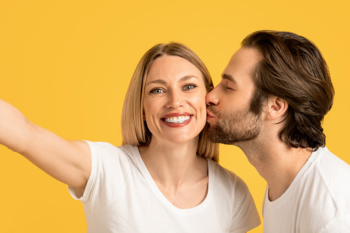 Happy millennial caucasian husband kissing wife on cheek in white t-shirt, lady make selfie, isolated on yellow background, close up. Photo for blog and social networks. Love, romance, relationship