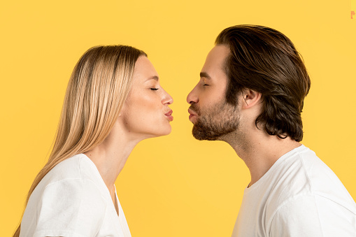 Cheerful millennial caucasian man and blonde lady in white t-shirts are kissing, isolated on yellow background, close up, profile. Couple relationship, love, romantic, ad and offer, facial expression