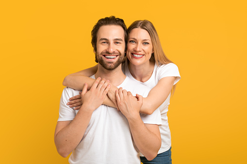 Cheerful millennial european female hugging from behind male with stubble in white t-shirts looking at camera, isolated on yellow background, studio, free space. Family love, relationship and romance