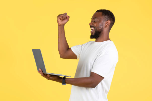 istock Cheerful Black Male Using Laptop Gesturing Yes Over Yellow Background 1475330260