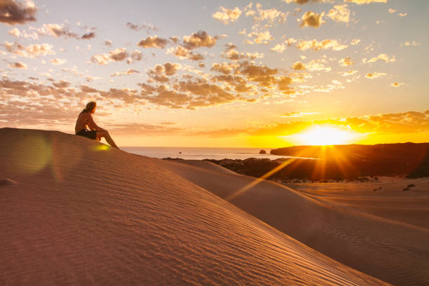 giovane che si siede sulla remota duna di sabbia arida del deserto dalla costa al tramonto - majestic landscape arid climate beach foto e immagini stock