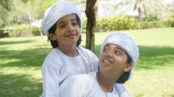 Emirati kids wearing traditional dress kandura playing together in a park. Happy children in white dress from United Arab Emirates
