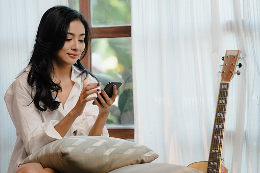 Smiling girl use a mobile phone to chatting online message or shopping online from home in the morning, Happy young asian woman relax on comfortable couch at home texting messaging on smartphone