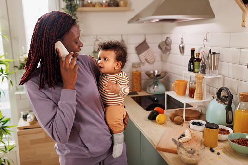 Mother talking on a phone while holding cute little baby boy in baby carrier at domestic kitchen