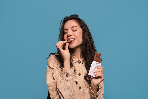 Glad happy millennial european woman with closed eyes enjoys taste of chocolate bar, isolated on blue background, studio shot, free space. Sweet tooth, love of sweets and candy, ad and good offer