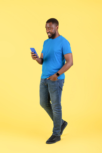Vertical Shot Of Black Guy Using Cellphone With Mobile App Standing In Studio Over Yellow Background. New Application Advertisement, Technology Concept. Full Length