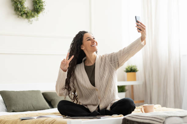 istock Young smiling woman sitting on bed taking selfie 1475343324