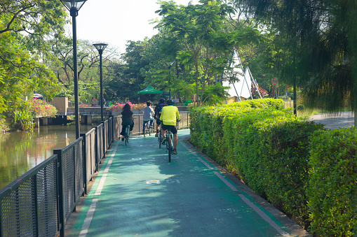 Rear view of cycling thai people  in Wachirabenchathat Park (Rot Fai Park) in Bangkok Chatuchak. People are cycling between fence to outer canal and a hedge with playground. Two men are wearing sports clothing and cycling helmets