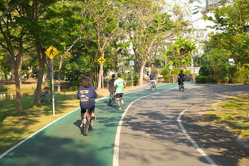 Cycling young thai women and a jogging man captured in Wachirabenchathat Park (Rot Fai Park) in Bangkok Chatuchak, Scene is at crossing between bicycle lane and pedestrian footpath. Jogger is crossing bicycle lane turning left. At left side is warning sign