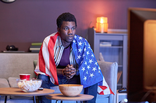 Portrait of young black man as sports fan watching match on Tv at home and wearing USA flag