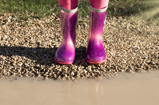 Child wearing new bright pink rain rubber boots standing near muddy puddle, no face. Preschool in wellies. Footwear for rainy weather