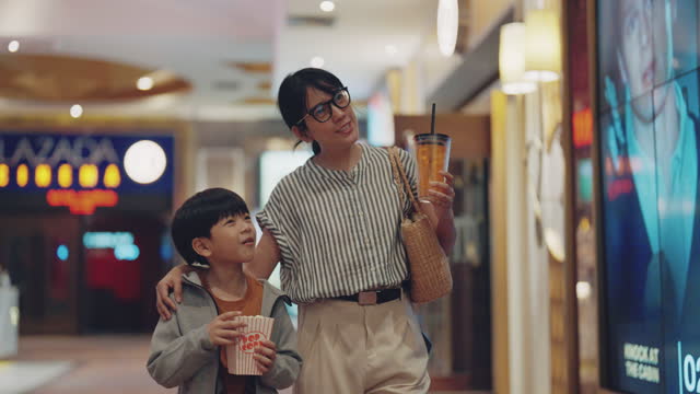 Modern asian woman and her son holing cup of fizzy drink and bucket of popcorn walking to cinema hall to watch movie.