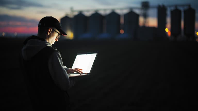 SLO MO Farmer works late on his laptop on the field with silos in the background,shot at blue hour