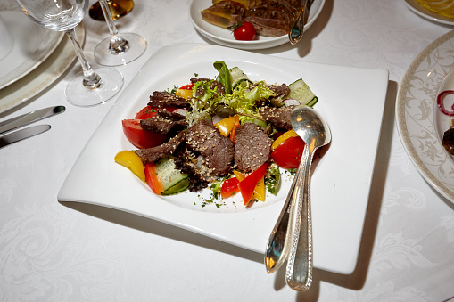 Sliced grilled beef steak with green leaves salad, cucumbers, tomatoes, bell peppers and sesame seeds on a white plate. Barbecue steak and healthy salad, top view. Table setting in a restaurant