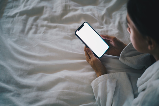 High angle view of a woman lying on the bed at night and looking at blank smartphone screen