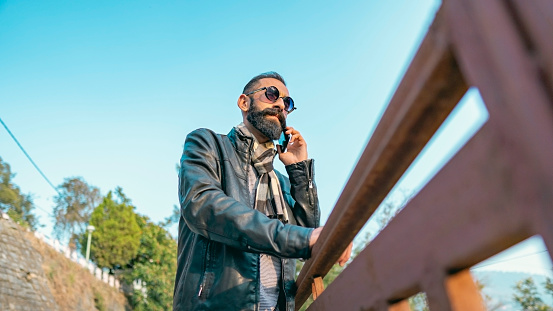 An Asian/Indian young man with a long beard uses a smartphone outdoors on a sunny day in winter. He wears round sunglasses and a  casual dress in this wide-angle image.