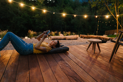 Photo of a young woman using mobile phone while relaxing on a wooden decking in front of a cabin she is renting for a vacation