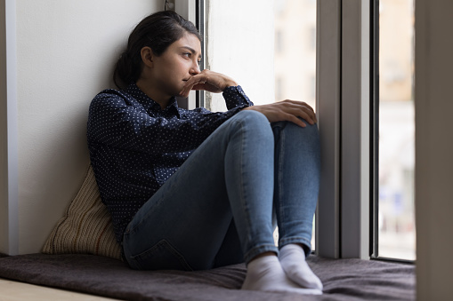 Depressed upset Indian woman resting on soft windowsill, looking out of window lost in bad negative thoughts, going through emotional crisis, depression, stress, feeling sad, frustrated