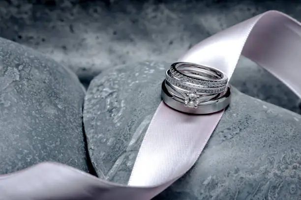 The bride and groom's wedding rings sit on top of a pink satin ribbon laying across grey stone carved hearts.