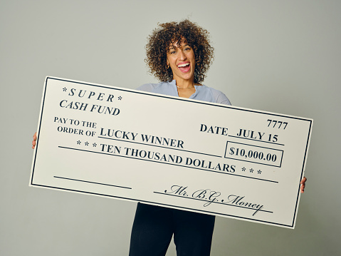 A happy young woman in a studio holding a giant prize check against a gray background.