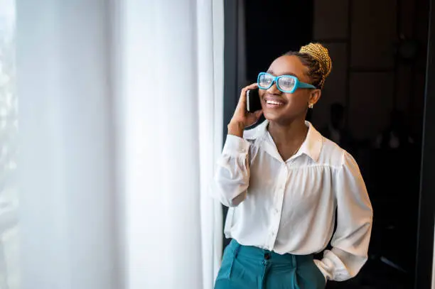 A beautiful black woman with blue eyeglasses and a bun in white shirt and dark green pants seen standing by the window and smiling while talking on the phone during a break between meetings.