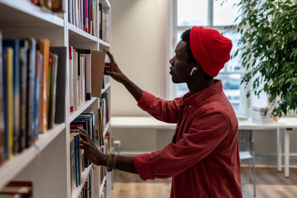 Thoughtful African American student man in red hat choosing book in college library or bookstore Thoughtful African American student man searching materials for educational research in college library. Young black hipster guy choosing book for reading in bookstore. Literature and education bookstore stock pictures, royalty-free photos & images