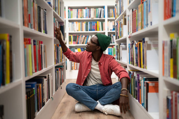 istock Young African American guy sits on floor in library between bookshelves, student searching for book 1475383697