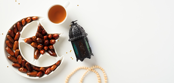 Ramadan Kareem banner design. Crescent moon plate with dried dates fruit, islamic lantern, cup of tea, rosary on white background.
