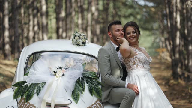 Stylish groom in a gray suit and a beautiful bride in a white lace dress smiling and hugging while sitting on an old timer car under the pine trees forest, slow motion
