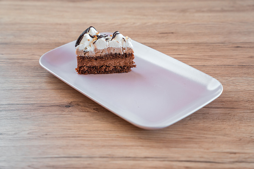 Professionally served slice of chocolate and nut cream cake, topped with abundance of whipped cream and dark chocolate. High angle view of cake on rectangular plate.