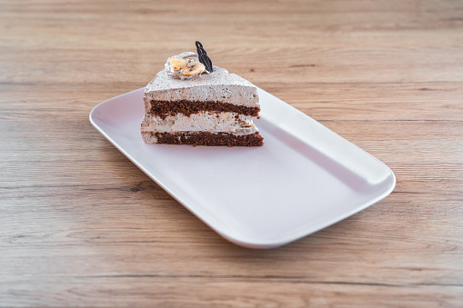 A slice of vegan almond and nuts cream cake, professionally decorated and served on a rectangular ceramic plate at a popular confectionery. Wooden surface under the plate, high angle shot. Full length image.