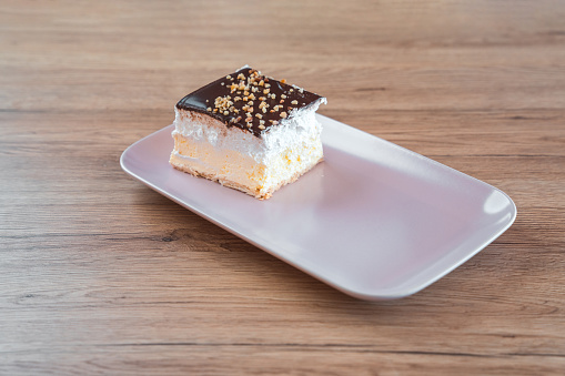 A slice of delicious creamy dessert displayed on a white, rectangular serving plate. Layers made of custard cream and puff dough, top is decorated with chocolate and biscuit crumbs. High angle view. Wooden shelf under. Full length image.