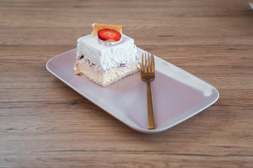 Slice of delicious creamy  dessert displayed on an elegant, rectangular serving plate. Layers of custard, white cream and puff dough visible from the side, top is decorated with a slice of strawberry and pieces of puff dough. High angle view. Fork next to it.