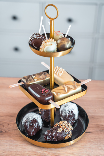 High angle view of delicious cake pops and other desserts on a three storey cake stand, all sweets covered in glossy chocolate fondant. Wooden surface under the cake stand.  Full length vertical image.