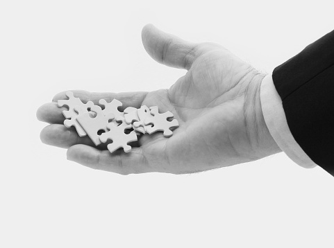 A man's hand holding white puzzle pieces, with part of the sleeve and professional business coat.