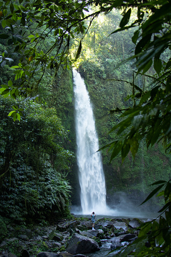 Young man on top of a rock taking a picture of a tall jungle waterfall in a lush rainforest - Bali, Indonesia