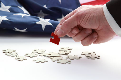 A man's hand holding a red puzzle pieces, with part of the sleeve and professional business coat, with American flag in background.