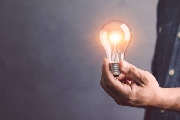 istock Businessman holding a clear light bulb with light effect on gray space wall background. Concept of new ideas for work. 1475418002