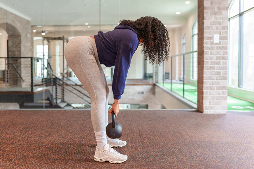 Profile view of a multiracial woman leaning down and doing deadlifts with a kettlebell during a functional fitness workout at the gym.