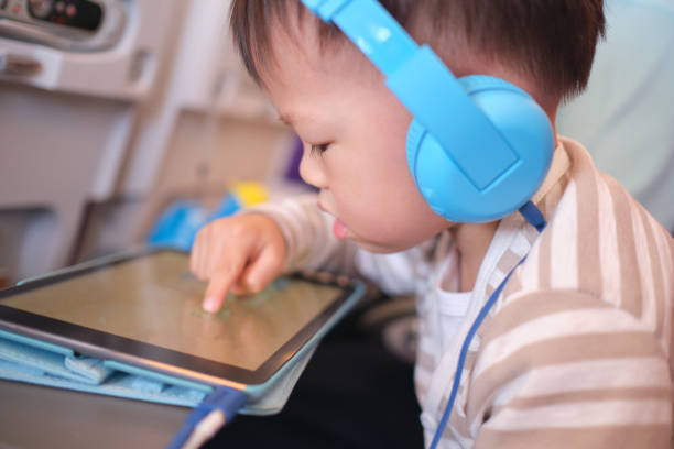 Asian toddler boy child wearing headphones using tablet pc watching cartoons, playing game during flight on airplane Cute Asian 3 years old toddler boy child wearing headphones using tablet pc watching cartoons, playing game during flight on airplane. Happy Flying with children concept, Soft and selective focus screen time and technology in babies stock pictures, royalty-free photos & images