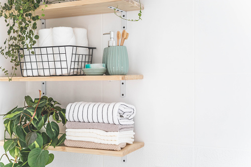 Beautiful white bathroom. Wooden shelves. Rolled towels, stacked towels and baskets. Soap, washcloth, toilet paper and toothbrushes. Indoor flowers. Eco style, minimalism.