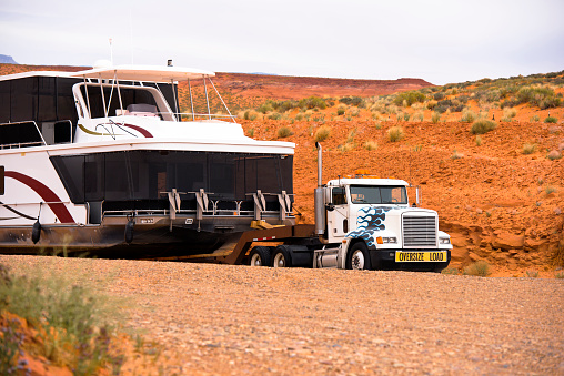 Big powerful American bonnet rig semi truck with sign oversize load transporting down the red rocks hill to the water a huge oversize multilevel ship on marine resort at Lake Powell in Page Arizona