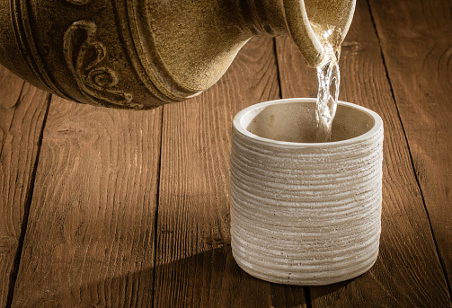 istock A jet of water pours from an earthenware jug into an earthenware glass standing on a wooden table 1475443502