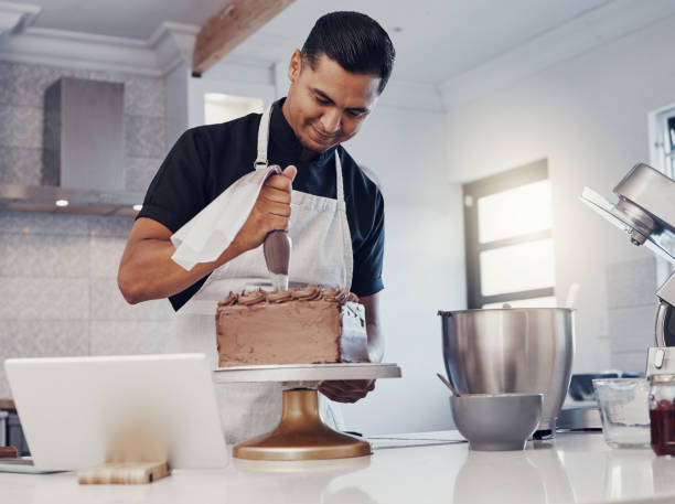 istock Piping, decorating and man baking a cake with chocolate in a kitchen or pastry chef happy with online recipe. Food, desert and cook preparing a sweet meal in Brazil and using a tablet 1475462654